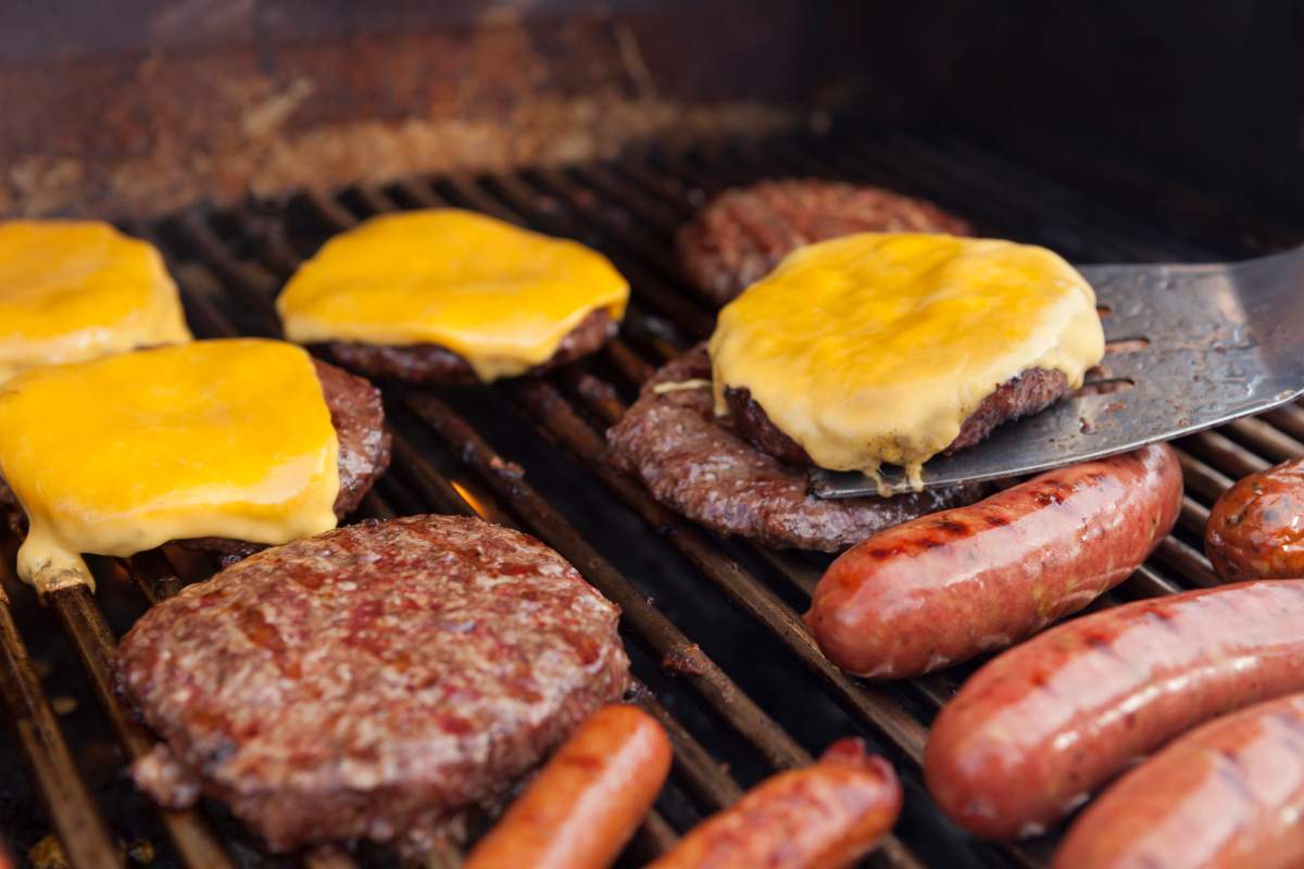 Outdoor photo of a grill with hotdogs, sausages, hamburgers and cheeseburgers. Shallow depth of field. Focus is on the cheeseburger and spatula.