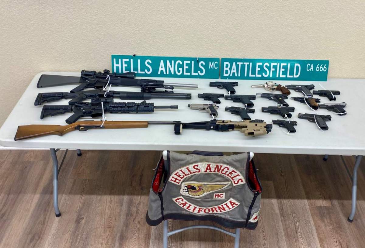 Several different types of guns sit on a plastic table. A Hells Angel vest is on a nearby chair.