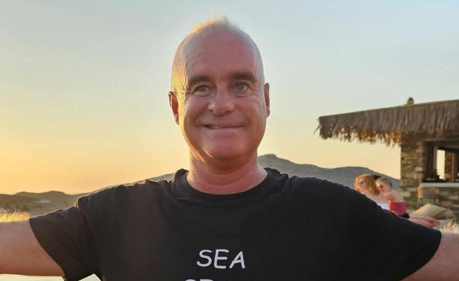 Eric Albert Calibet in a black T-shirt that says 'Sea.' He is smiling in front of a sunset.