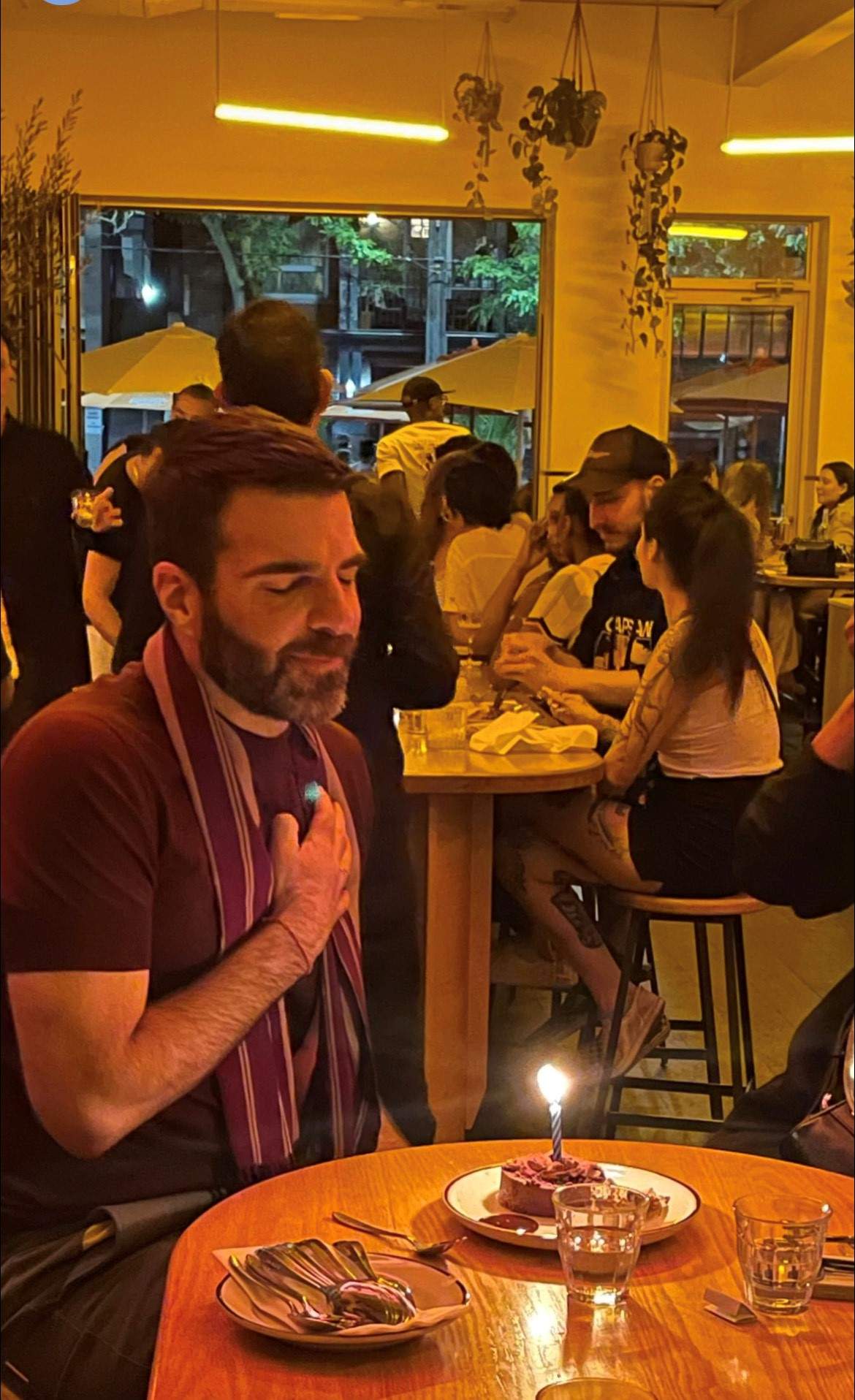 Zachary Quinto sitting in front of a slice of cake with a candle. His eyes are closed and his hand is on his heart.