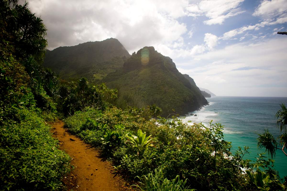 File photo of the Kalalau Trail, which snakes along the high sea cliffs of the Na Pali Coast. The photo features a dirt trail, lush green mountains and blue water.