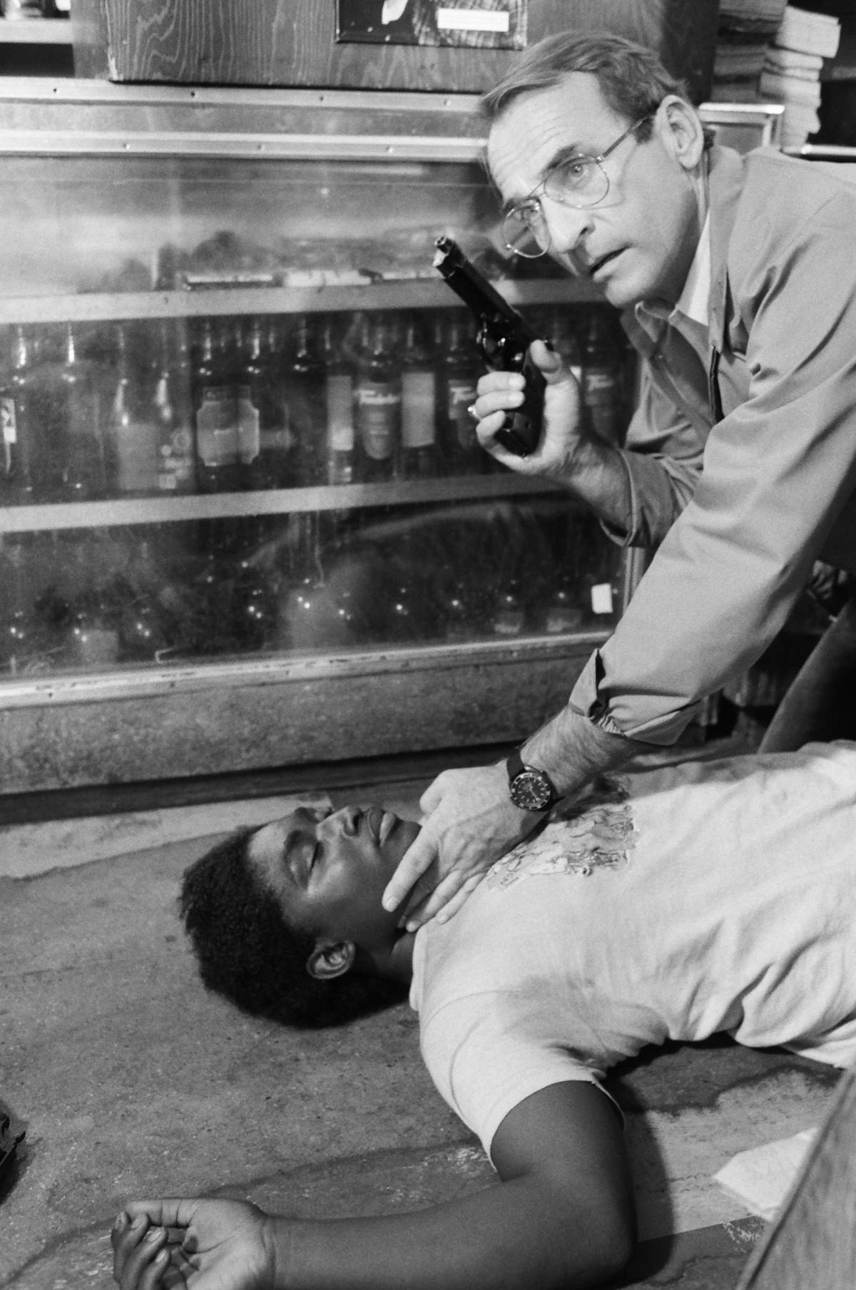 A black and white still from 'Hill Street Blues.' Sikking is holding a gun, checking a man's pulse as he lies on the ground.