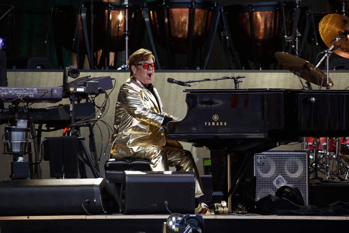 Elton John wearing a shiny gold outfit, playing a piano and singing into a microphone on a stage.