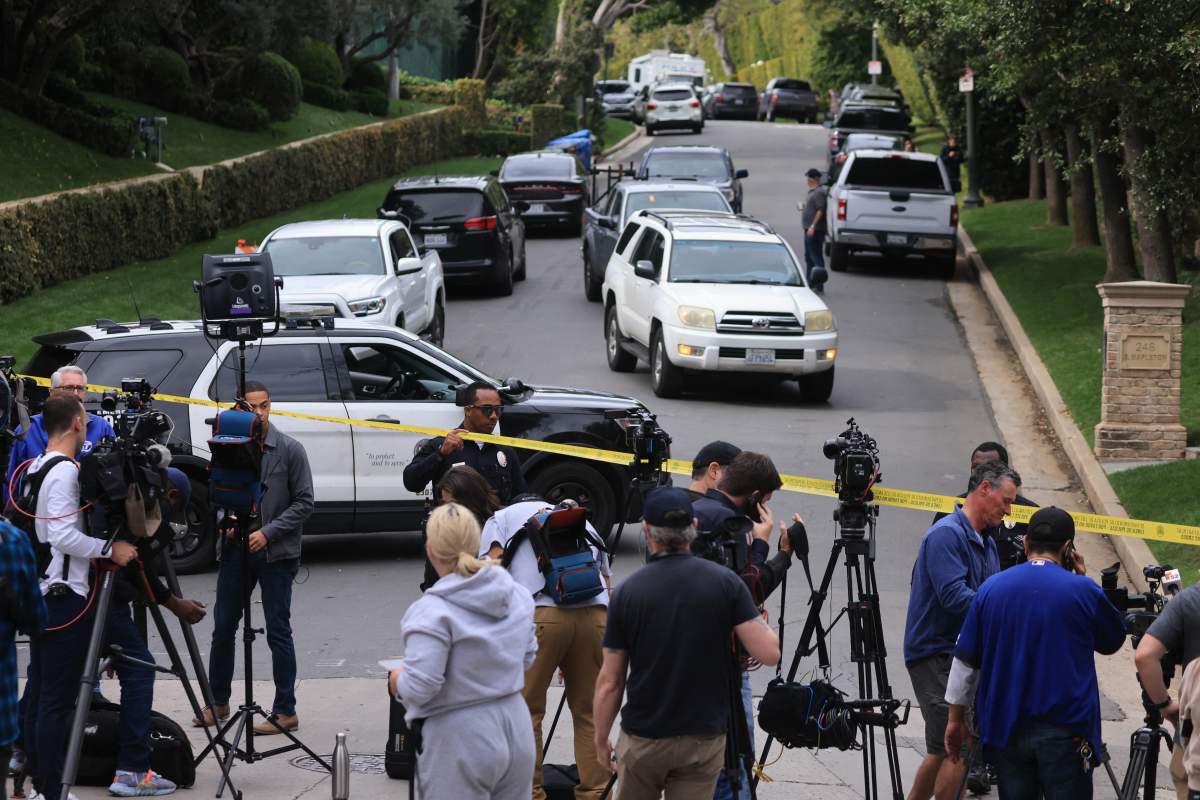 Police and press with cameras stand on a taped off road filled with cars.