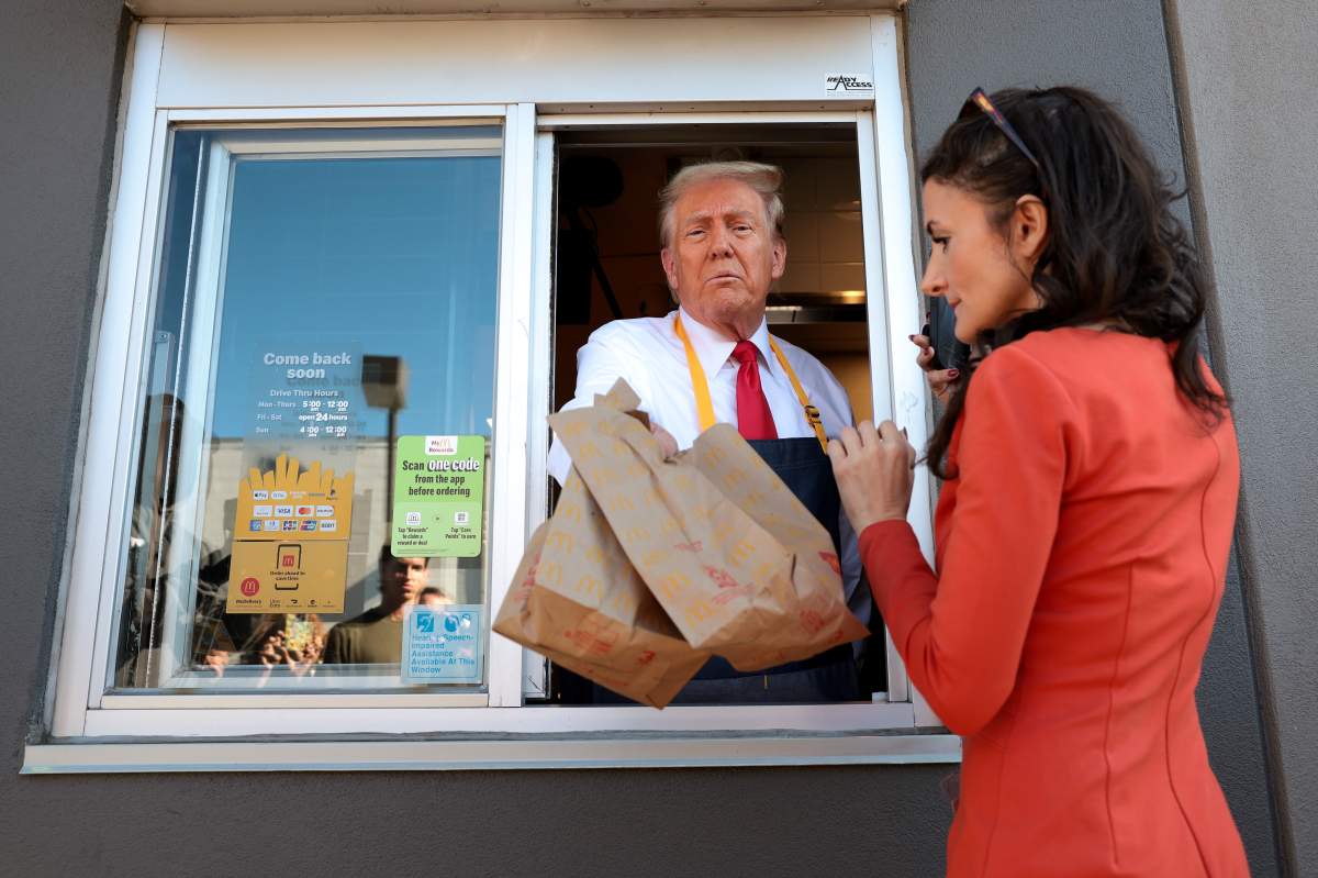Donald Trump in a McDonald's drive thru window. He is holding two brown bags, and another woman is in the frame.