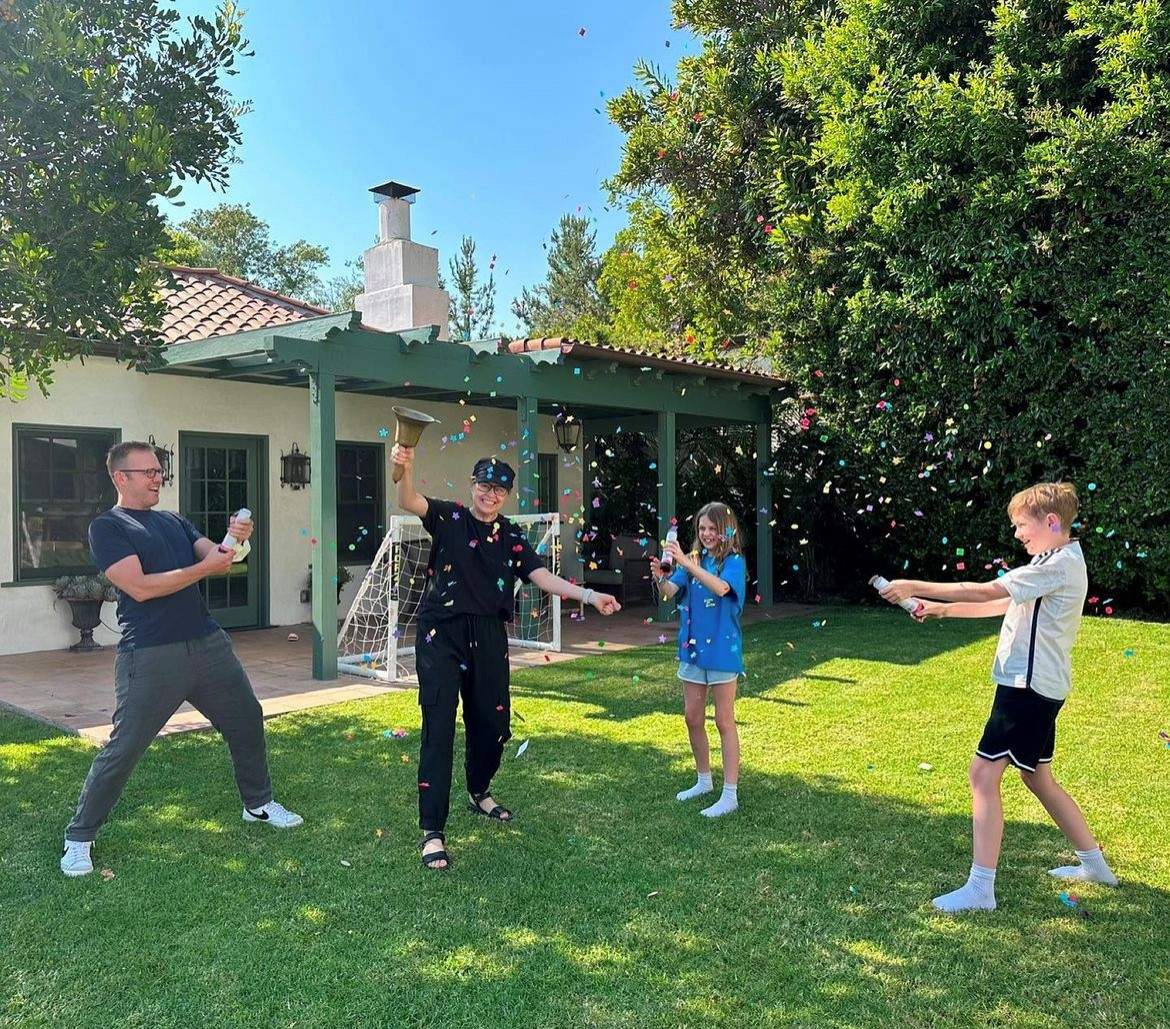 Jenna Fischer rings a bell in a backyard beside her husband, Lee, and their two children, who are throwing confetti.