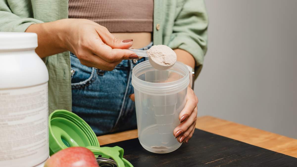 Woman in jeans and shirt with a measuring spoon in her hand puts portion of whey protein powder into a shaker on wooden table with white capsules, bananas and apple. Process of making protein drink.