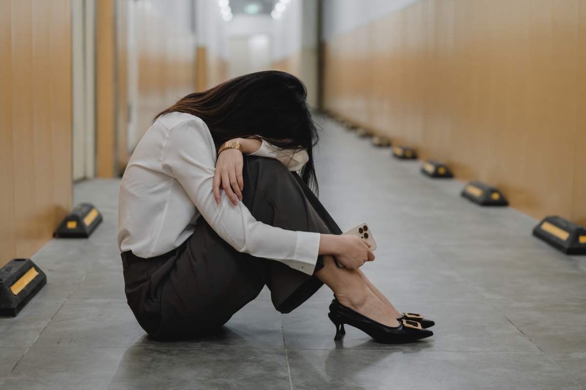 A businesswoman sits in the middle of a hallway with her face buried into her arm.