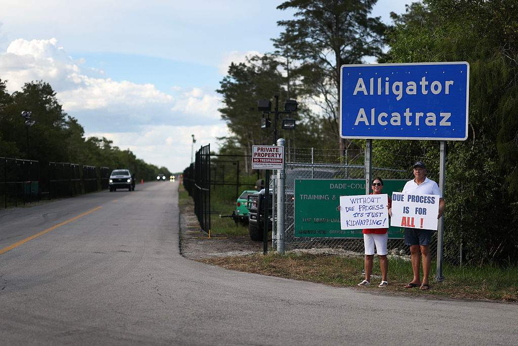 Leo and Catherine Gentile hold signs near the entrance to the state-managed immigration detention center dubbed Alligator Alcatraz, located at the Dade-Collier Training and Transition Airport in the Florida Everglades on August 03, 2025 in Ochopee, Florida.