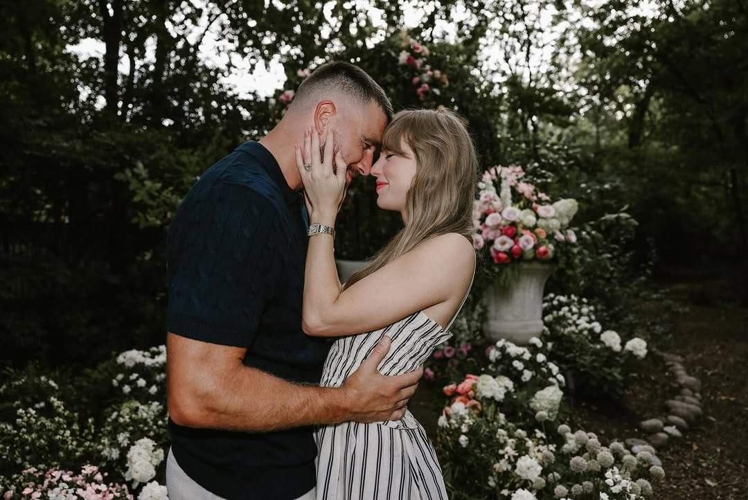 Taylor Swift, in a white dress with black stripes, holds Travis Kelce’s face. Kelce, dressed in a dark shirt, smiles at her with his eyes closed.