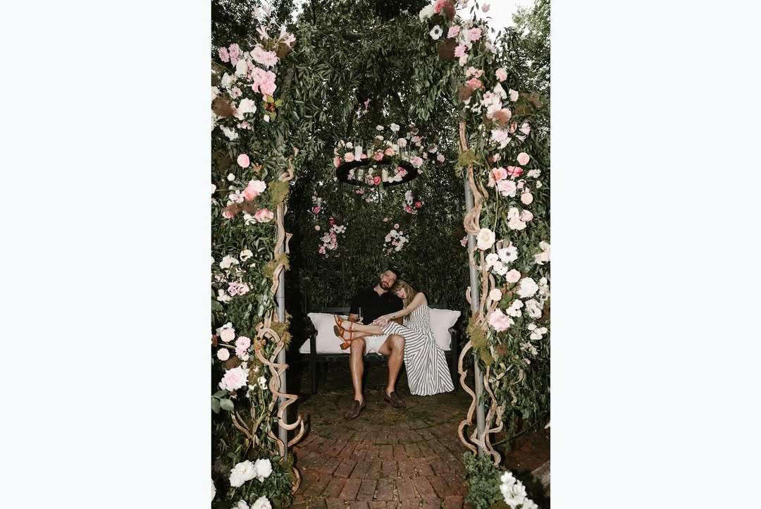 The last picture in the slide show is of the couple sitting on a bench beneath an archway full of flowers on trellises. Swift is resting her head on Kelce’s shoulder, with her legs across his lap. She wears brown sandals and Kelce is dressed in a pair of white shorts.