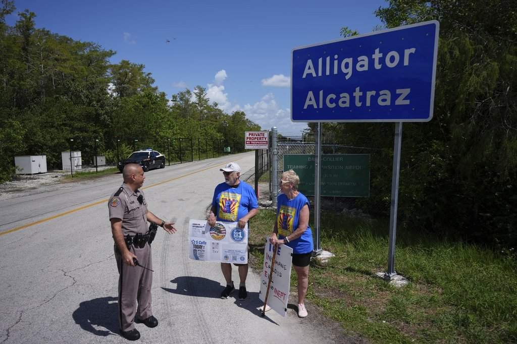 A police officer urges Art Sennholtz, 80, center, and Christy Howard, 70, of Just Us Volusia to be careful of fast-moving traffic as they hold protest signs outside the entrance to an immigration detention center in the Florida Everglades known as 'Alligator Alcatraz,' Thursday, Aug. 28, 2025, in Collier County, Fla.