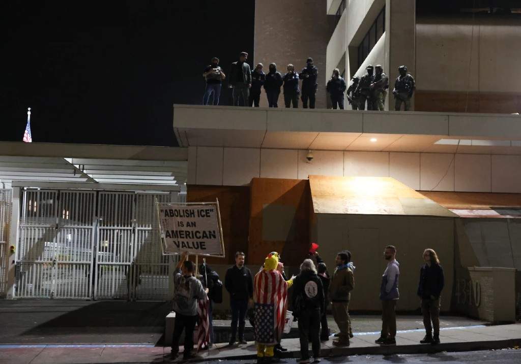People protest outside a U.S. Immigration and Customs Enforcement facility as federal agents watch from the rooftop, in Portland, Oregon, Wednesday, Oct. 1, 2025.