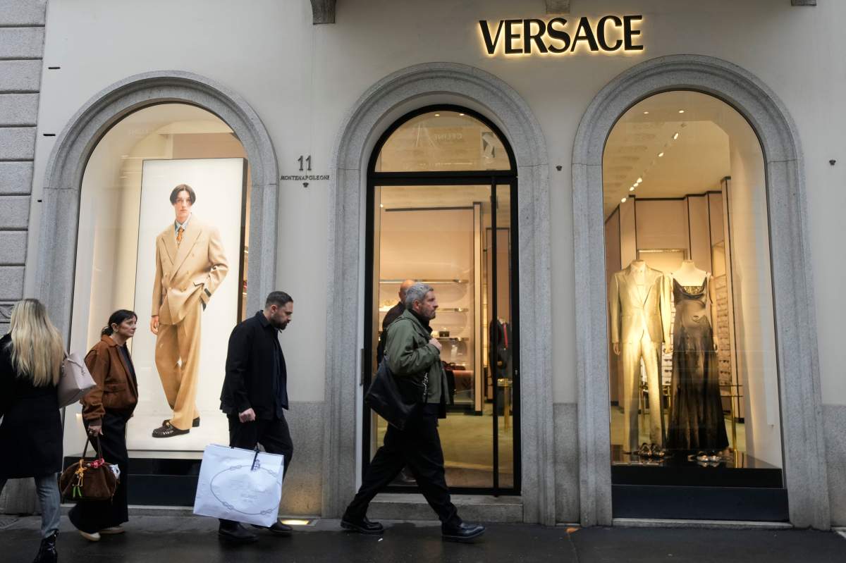 A man walks with a Prada shopping bag in front of a Versace shop, at the Montenapoleone luxury fashion street, in Milan, Italy, Tuesday, Dec. 2, 2025.