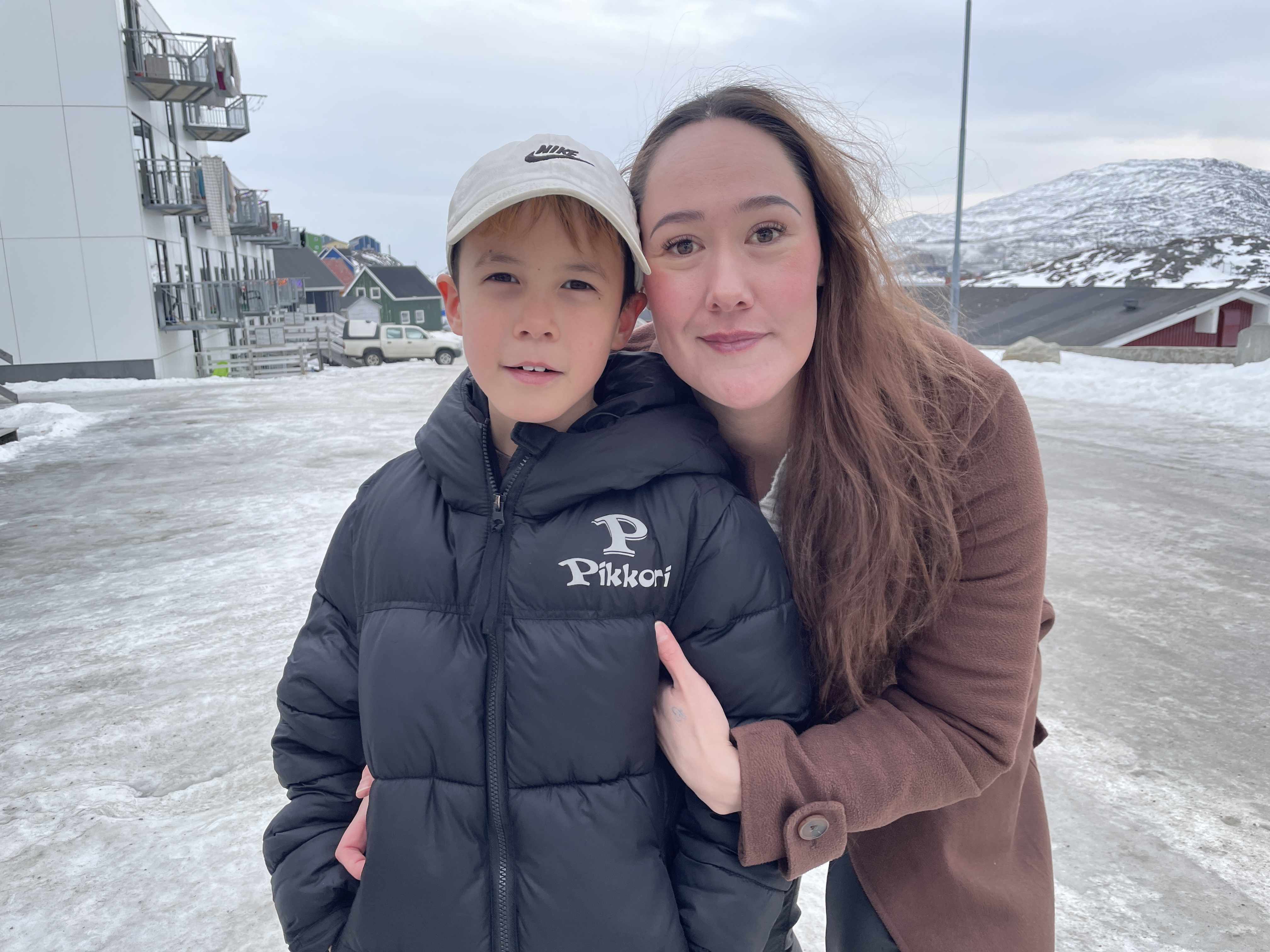 A woman with long brown hair wearing a brown coats stands behind a boy in a black puffer vest and white cap. They are outside on a snowy road.
