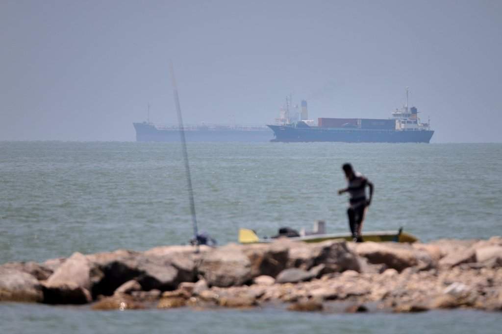 A man stands beside a fishing pole along the shore as cargo ships and commercial vessels are seen in the Strait of Hormuz off Bandar Abbas, Iran, Wednesday, June 17, 2026. (Amirhosein Khorgooi/ISNA via AP).