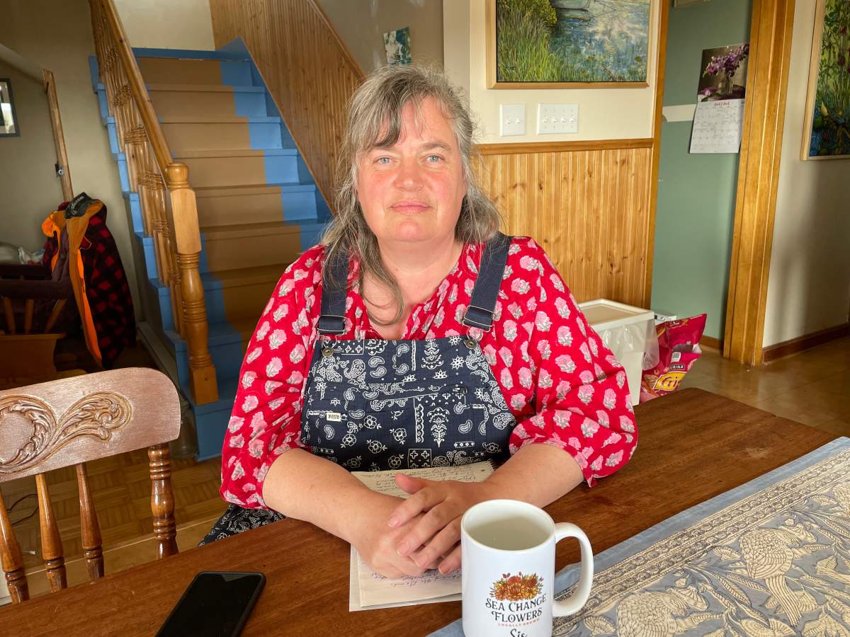 A photo of a woman with long grey hair and a bright red shirt sits at a kitchen table.