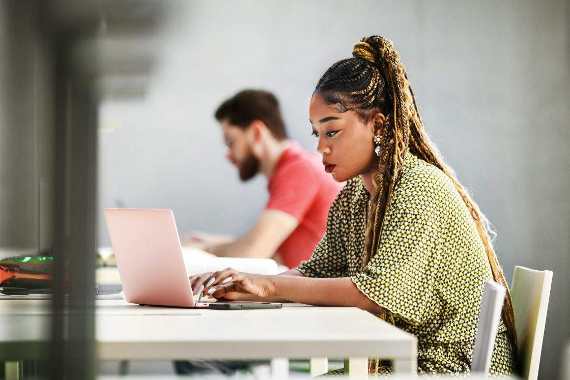 A young female student concentrates intently on her laptop while seated at a library.