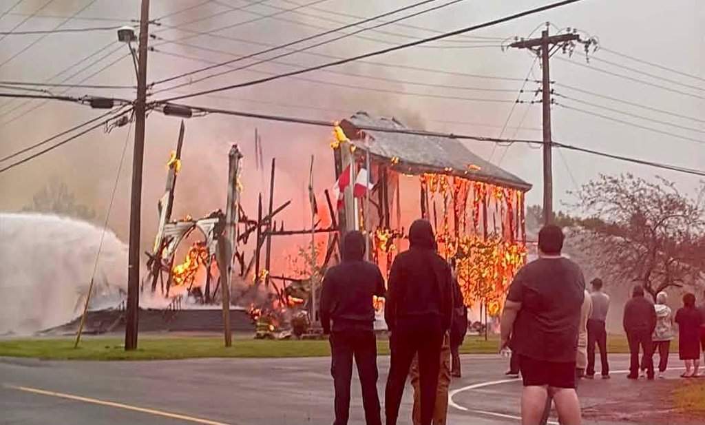 People look on as Saint-Simon Catholic Church in Saint-Simon, N.B., engulfed in flames on Monday Aug. 24, 2026. THE CANADIAN PRESS/Handout- Yvonne Chiasson (mandatory credit).