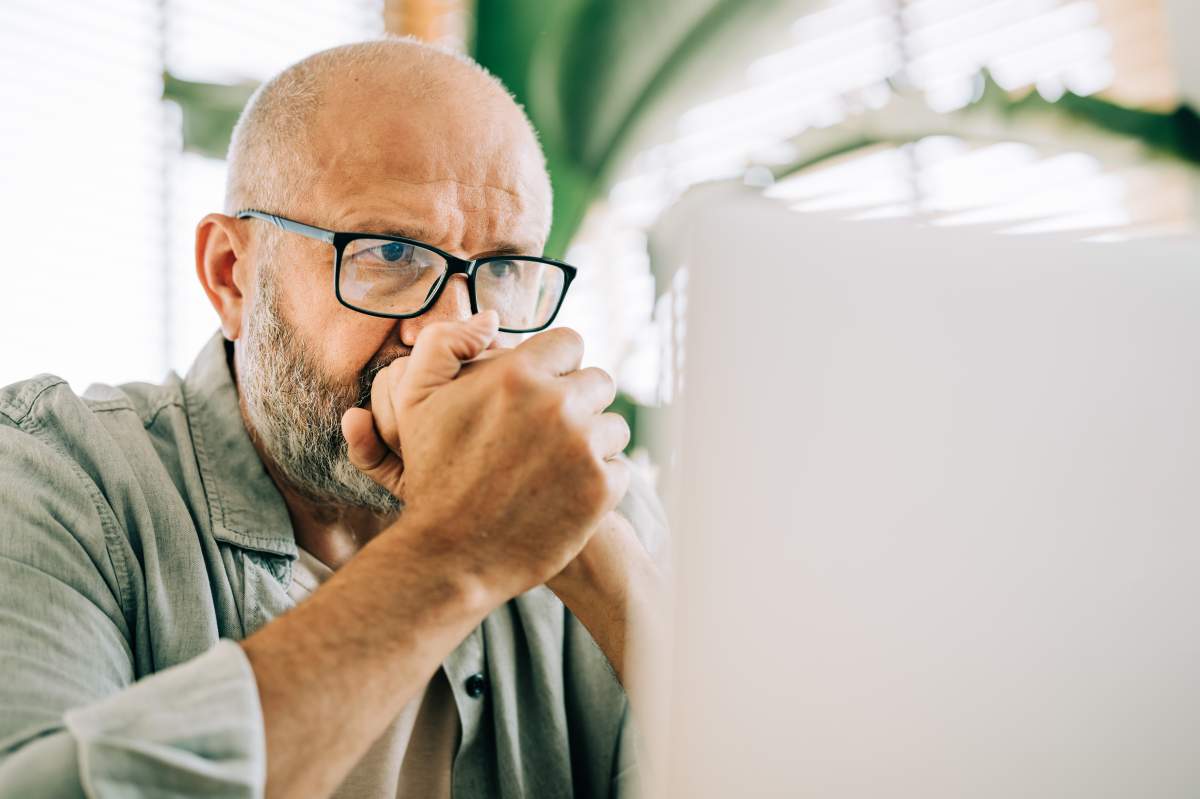 A close-up portrait of a thoughtful middle-aged man wearing glasses, looking intently at his laptop screen. His concentrated expression and clasped hands convey a sense of deep concentration and serious thought.