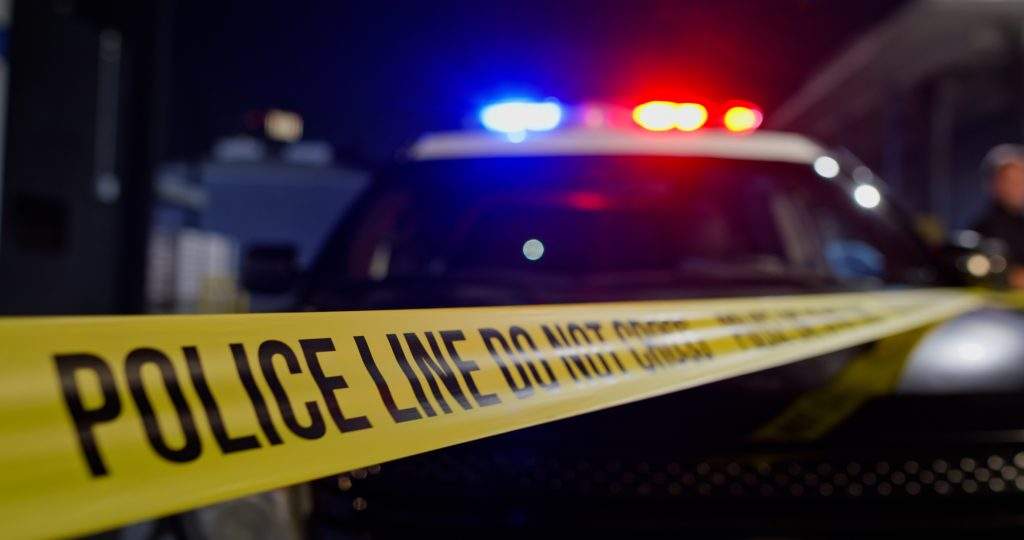 A police crime scene at night with yellow police tape in the foreground reading 'POLICE LINE DO NOT CROSS' .