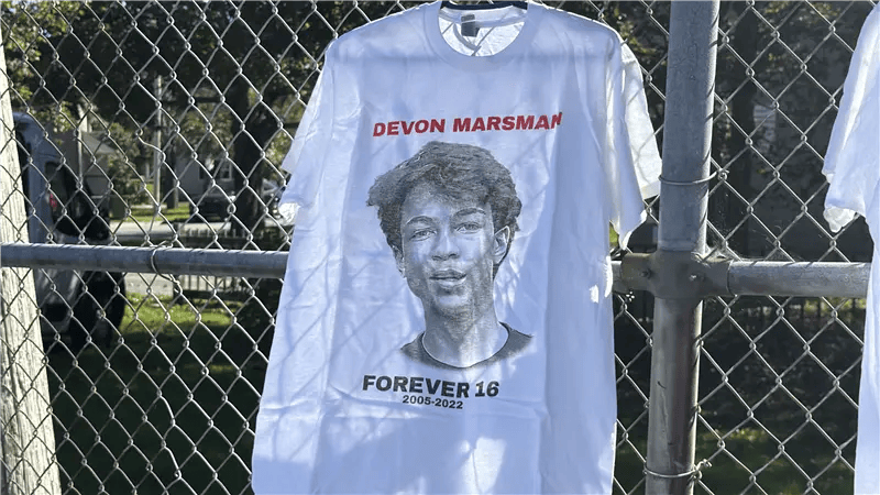 A white T-shirt hangs on the basketball court's fence. It has "Devon Marsman" written across the top in red letters, with Devon's face in black and white underneath. Below his photo it says "Forever 16."