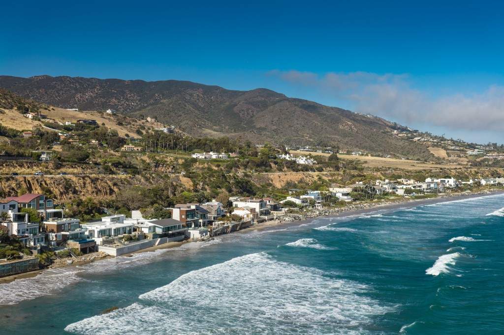 Aerial photograph of beachfront homes lining the shore near Malibu, California, with the Santa Monica Mountains rising in the background.